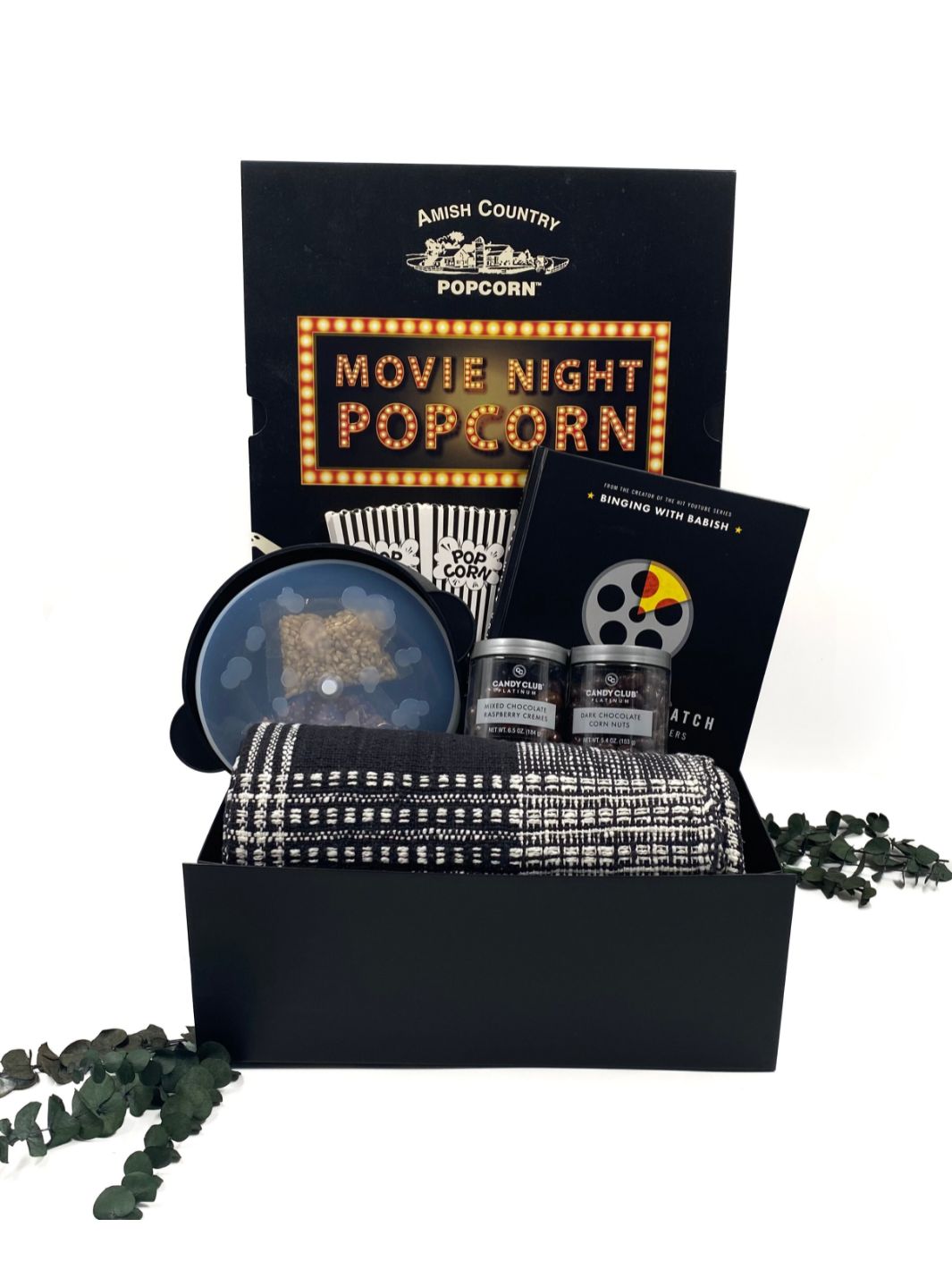 Open gift box displaying its contents, including a cozy black-and-white plaid throw, Eat What You Watch cookbook, silicone microwave popcorn popper, Movie Night Popcorn, mini popcorn bags, and gourmet chocolate treats.